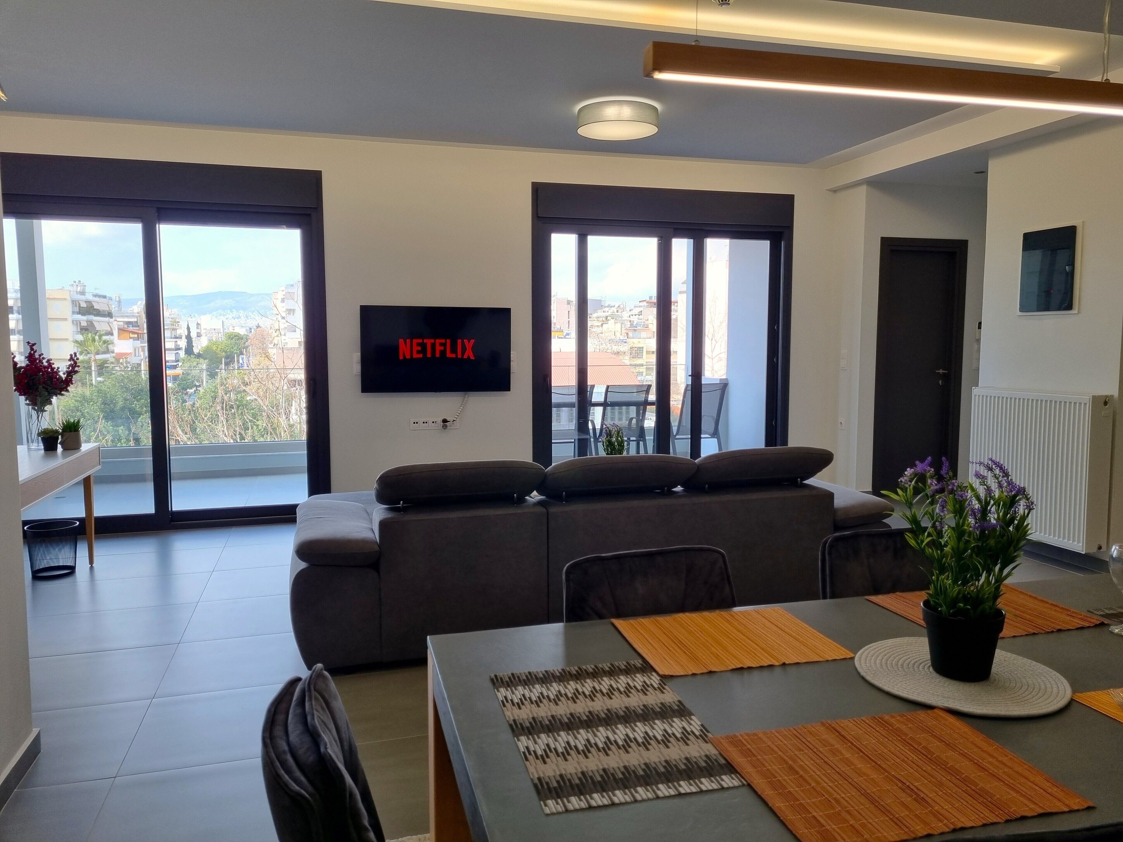 A modern living room with a sofa, a dining table with placemats, a TV mounted on the wall, and large windows offering a city view.