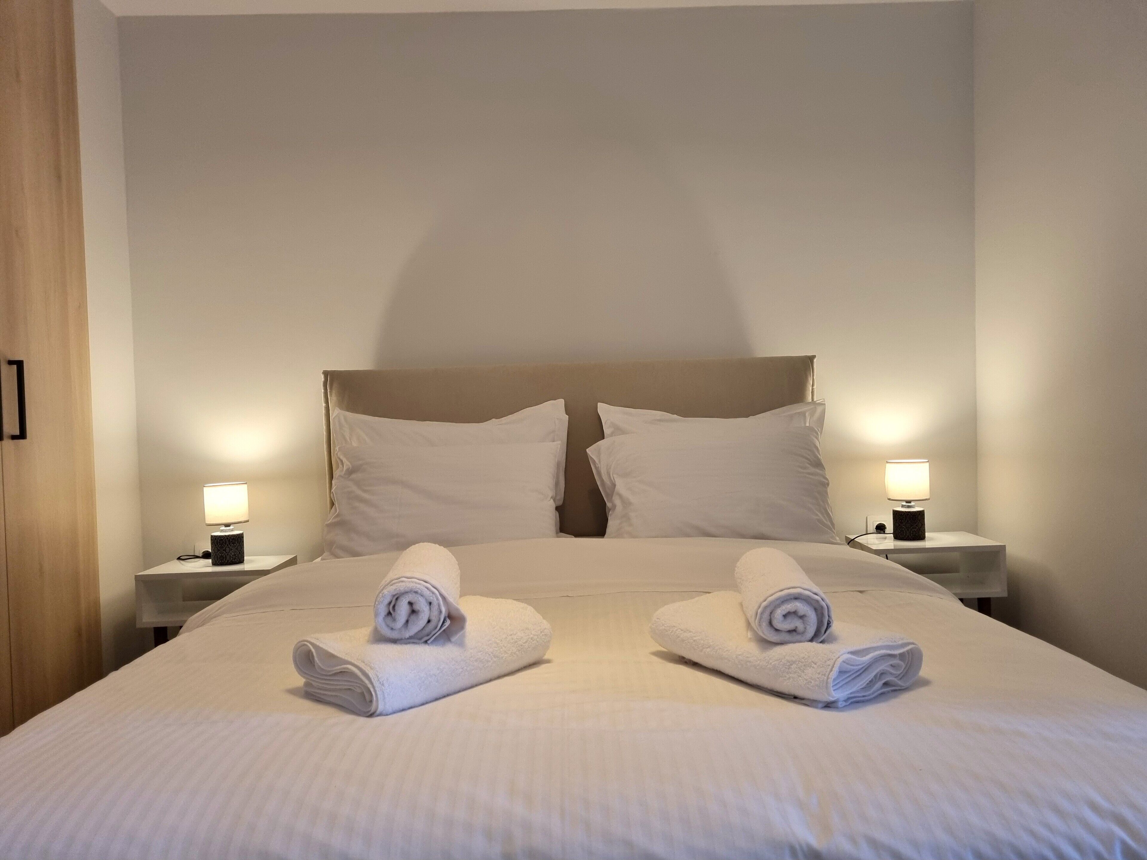 A neatly made bed with two pillows and two rolled towels placed on the bed. There are two bedside tables with lamps on each side of the bed.