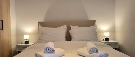 A neatly made bed with two pillows and two rolled towels placed on the bed. There are two bedside tables with lamps on each side of the bed.