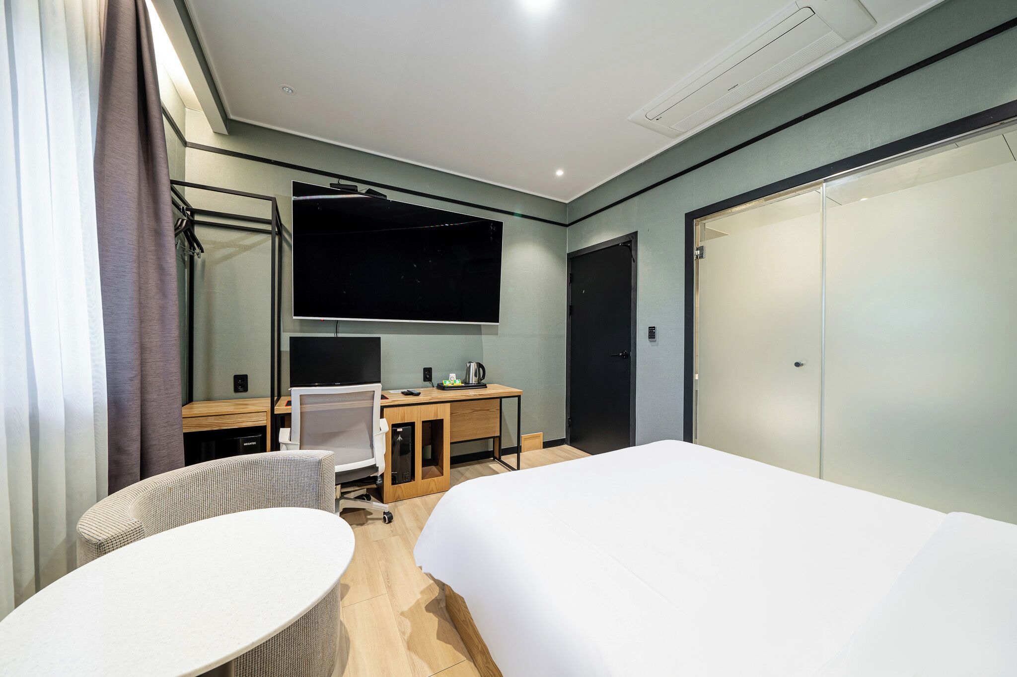 A modern hotel room with a bed, a desk with a computer, a chair, a TV, and a wardrobe.
