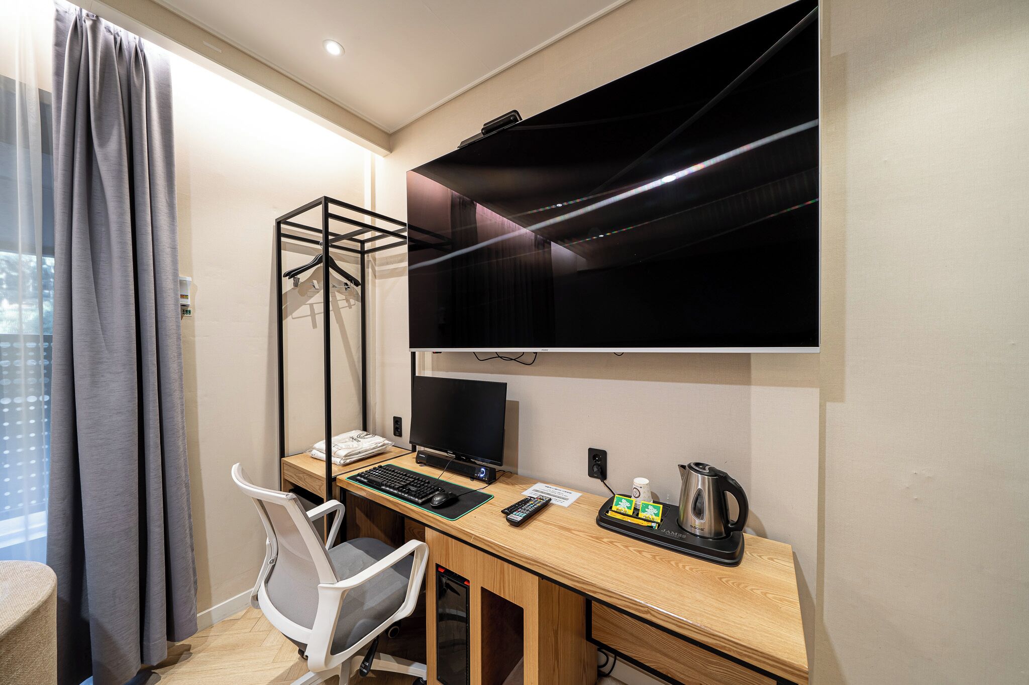 Habitación de hotel con escritorio, una silla, televisor montado en la pared, perchero y ventana con cortinas.