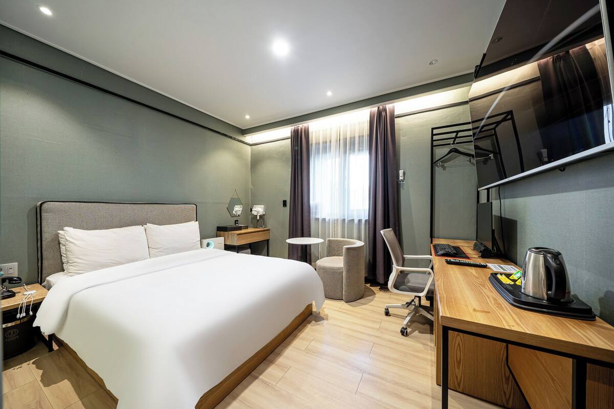 A hotel room with a large bed, a desk with a kettle, a chair, a small table, and a TV.