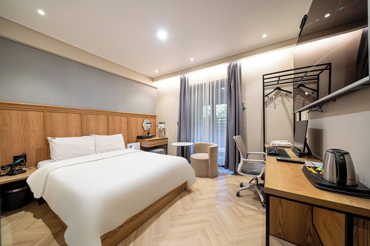 A hotel room with a large bed, a desk with a computer, a chair, a small table, and a mirror.
