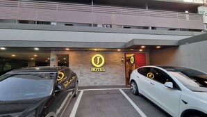 Entrada de hotel con un cartel, dos coches aparcados en la parte delantera y una zona de estacionamiento techada.