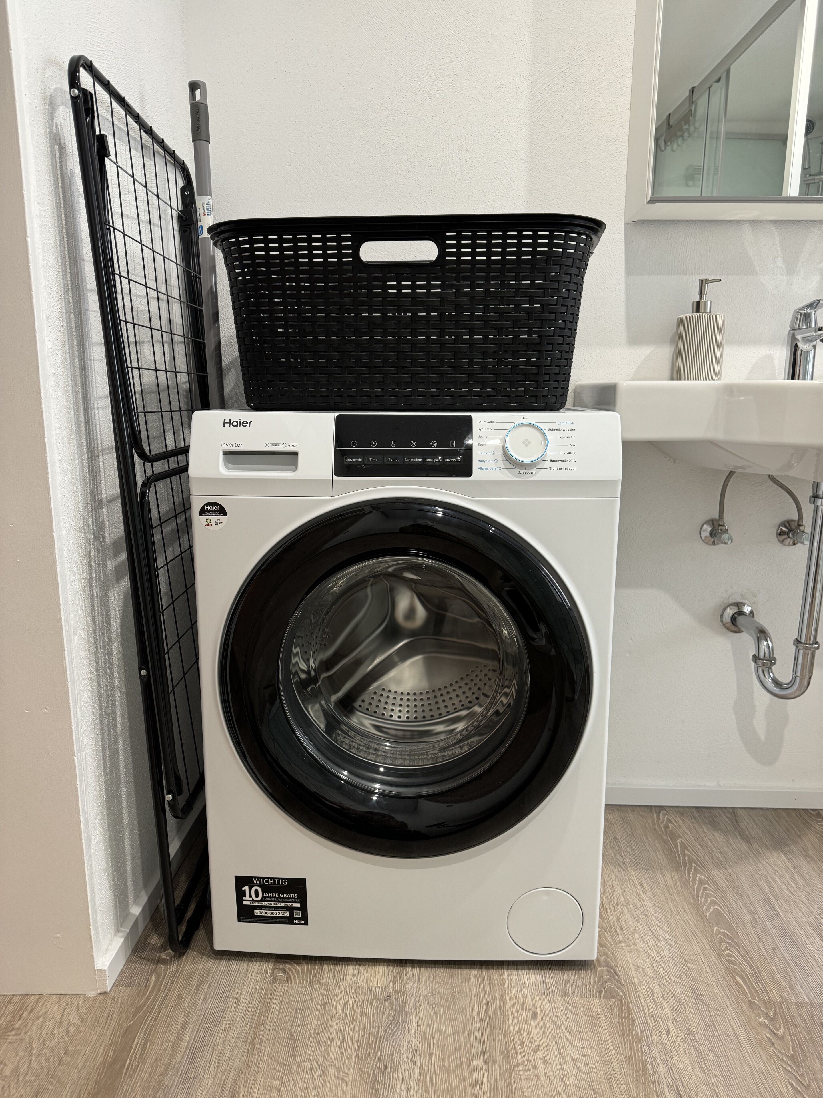 A front-loading Haier washing machine with a black basket on top, located in a room with a white sink and a mirror above it.
