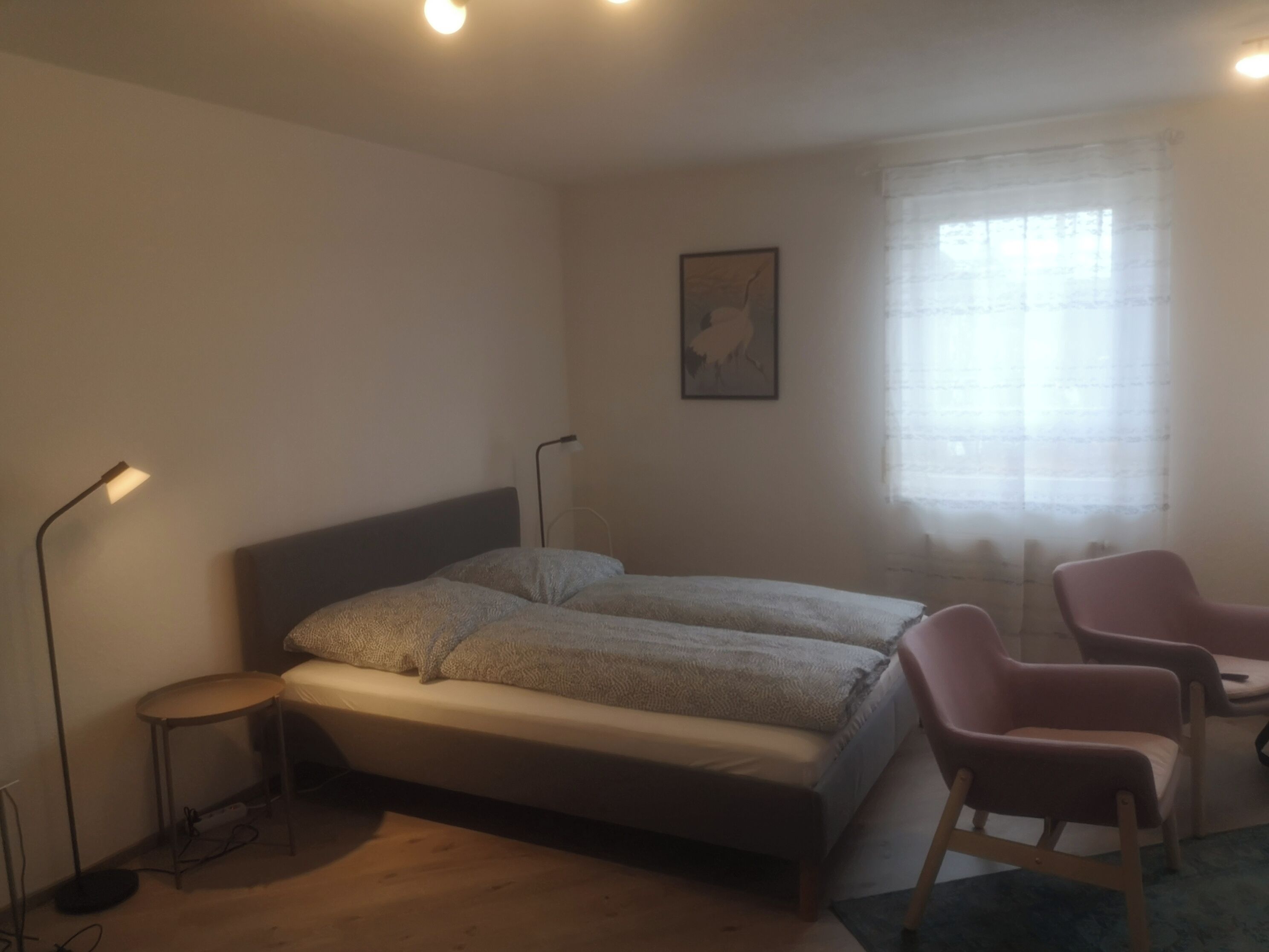 A bedroom with a bed, two bedside lamps, a chair, a small table, and a window with sheer curtains.