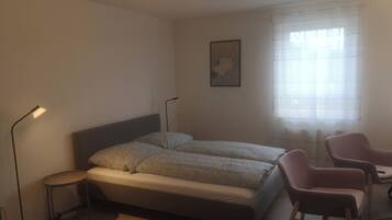 A bedroom with a bed, two bedside lamps, a chair, a small table, and a window with sheer curtains.