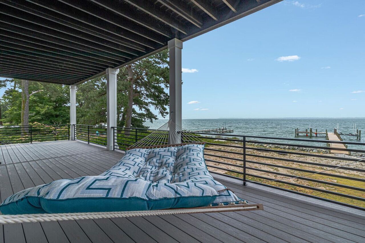 Une terrasse en bois avec un hamac et des coussins à motifs, donnant sur une plage et la mer.