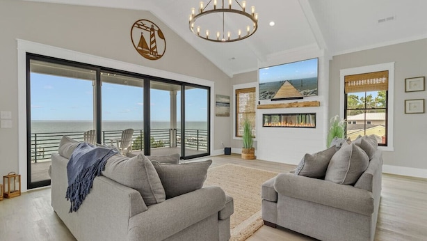 Ein geräumiges Wohnzimmer mit einer großen Schiebeglastür, die auf einen Balkon mit Meerblick führt. Zwei graue Sofas und eine Chandelier sind vorhanden.