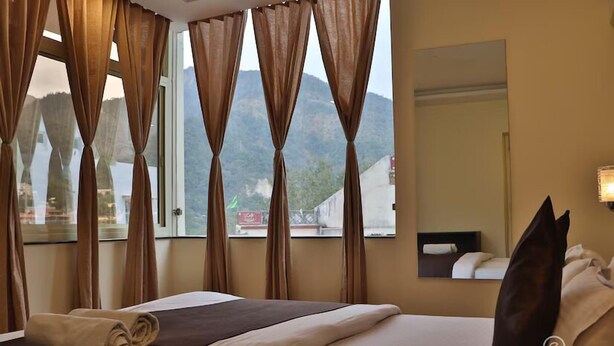 A hotel room with a bed, curtains, a window with a mountain view, and a mirror. - Shiv Shakti Hostel (Rishikesh)