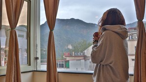 A person standing by a window with curtains, looking out at a mountainous landscape. - Shiv Shakti Hostel (Rishikesh)
