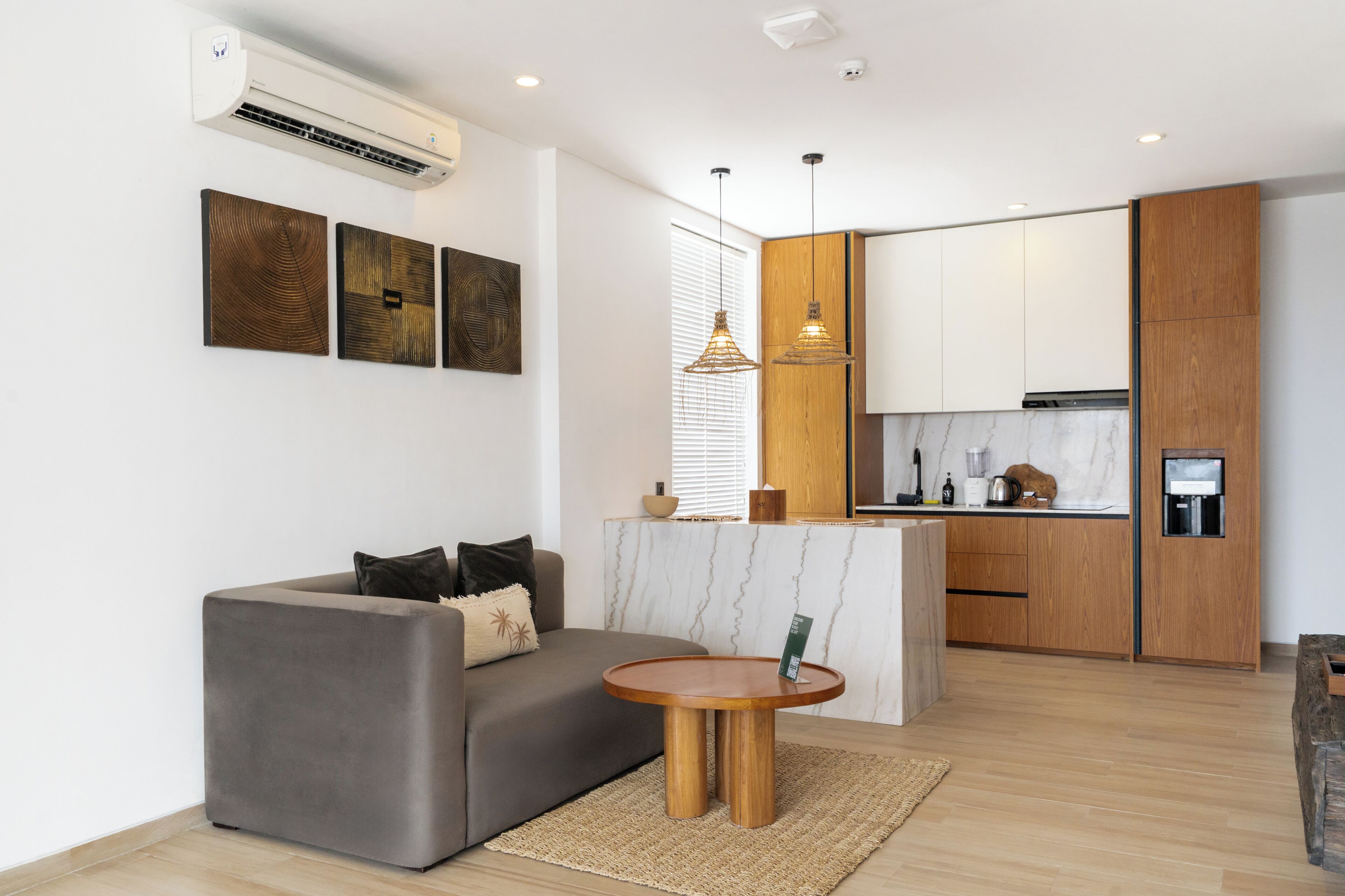 A modern living room with a sofa, a round wooden coffee table, and a kitchen area with wooden cabinets and a marble countertop.