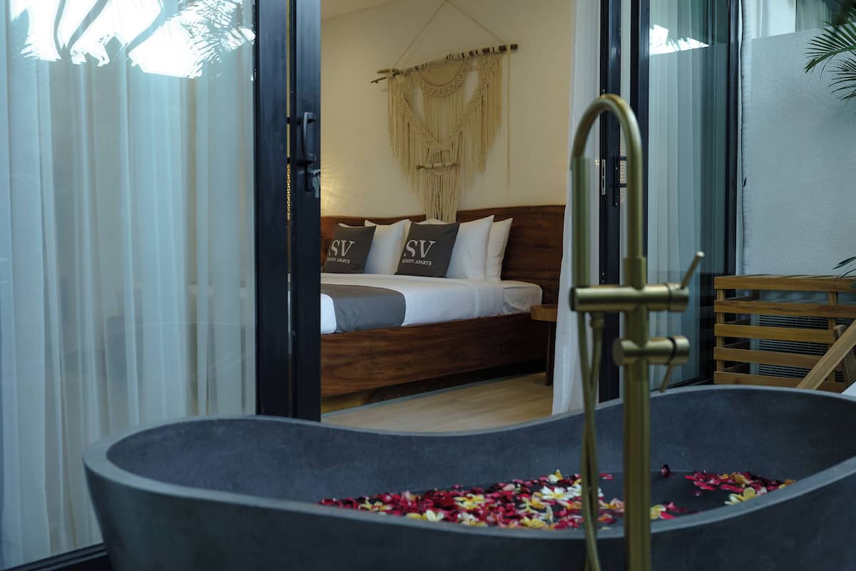 A bedroom with a bed, a macramé wall hanging, and a bathtub filled with flower petals.