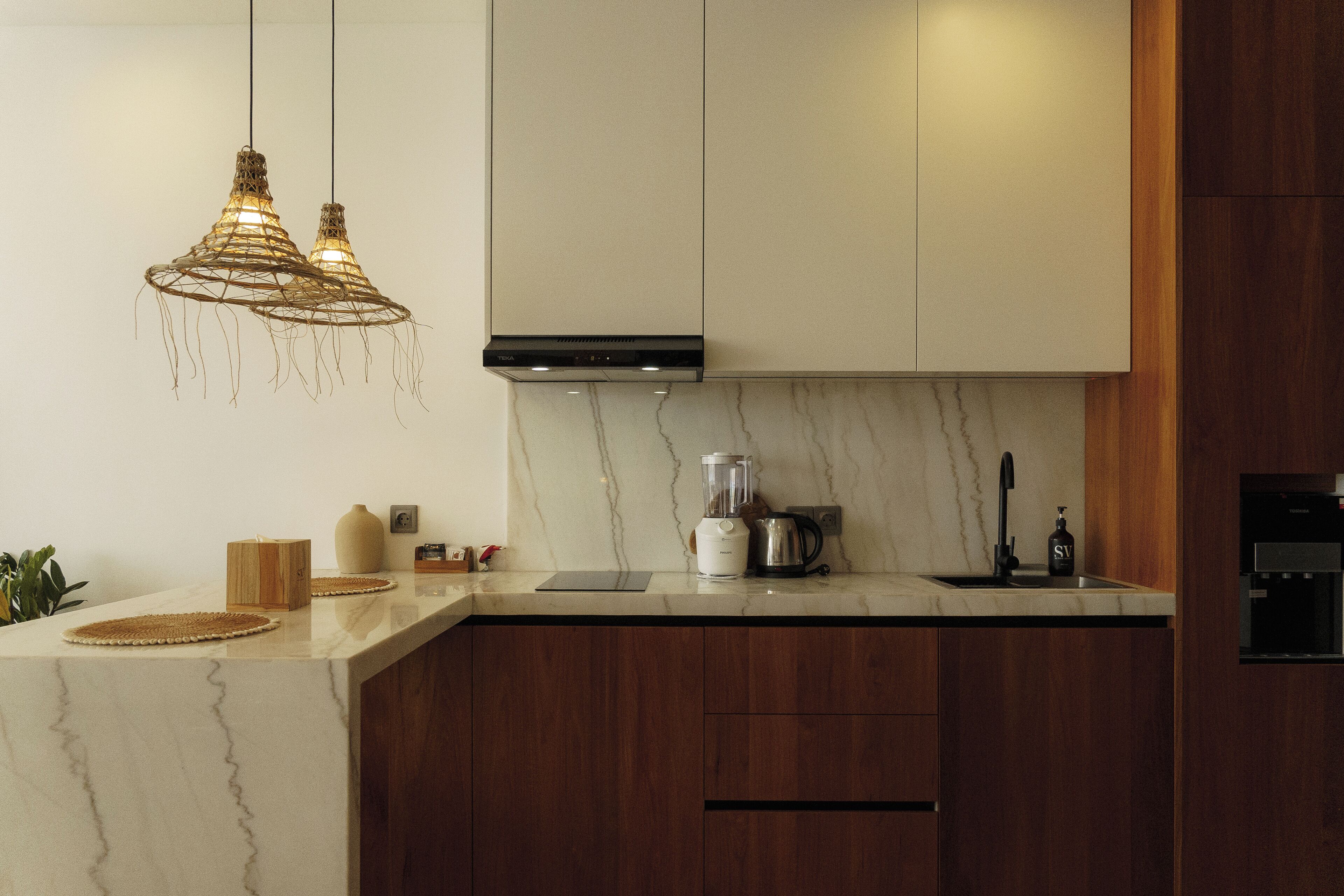 Een moderne keuken met een marmeren aanrechtblad, houten kastjes, een ingebouwde kookplaat en een hanglamp.