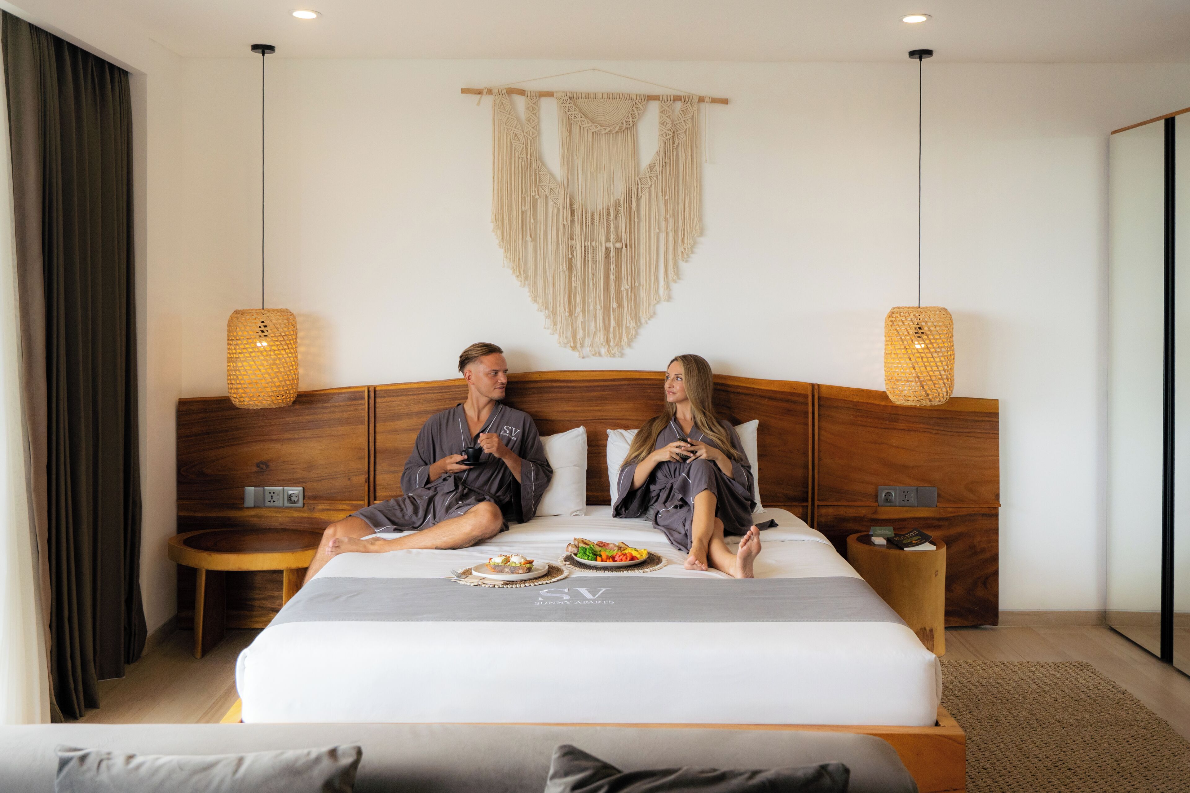 A modern hotel room with a wooden headboard, a macrame wall hanging, and a bedside table with a lamp.