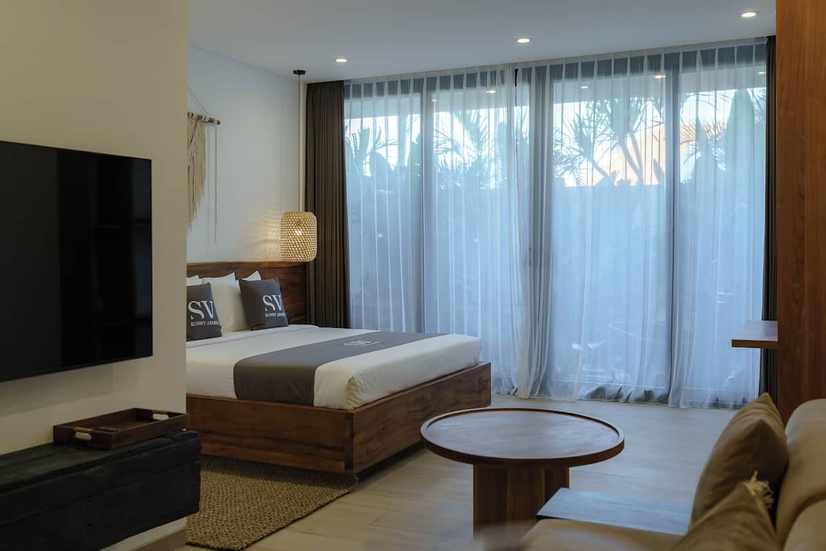 A modern hotel room with a large bed, a flat-screen TV, a wooden coffee table, and a view of palm trees through sheer curtains.