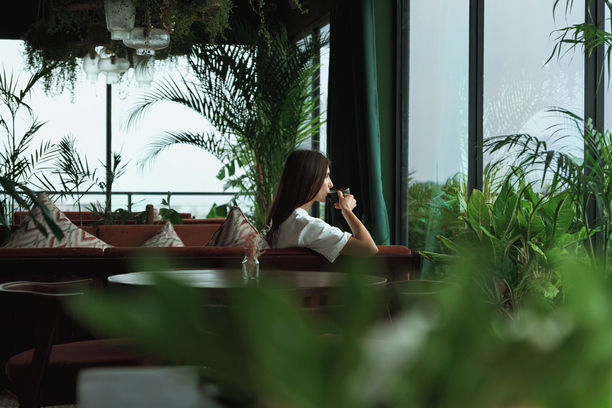 Una persona sentada en un sofá rojo en una habitación con ventanas amplias, rodeada de plantas verdes.