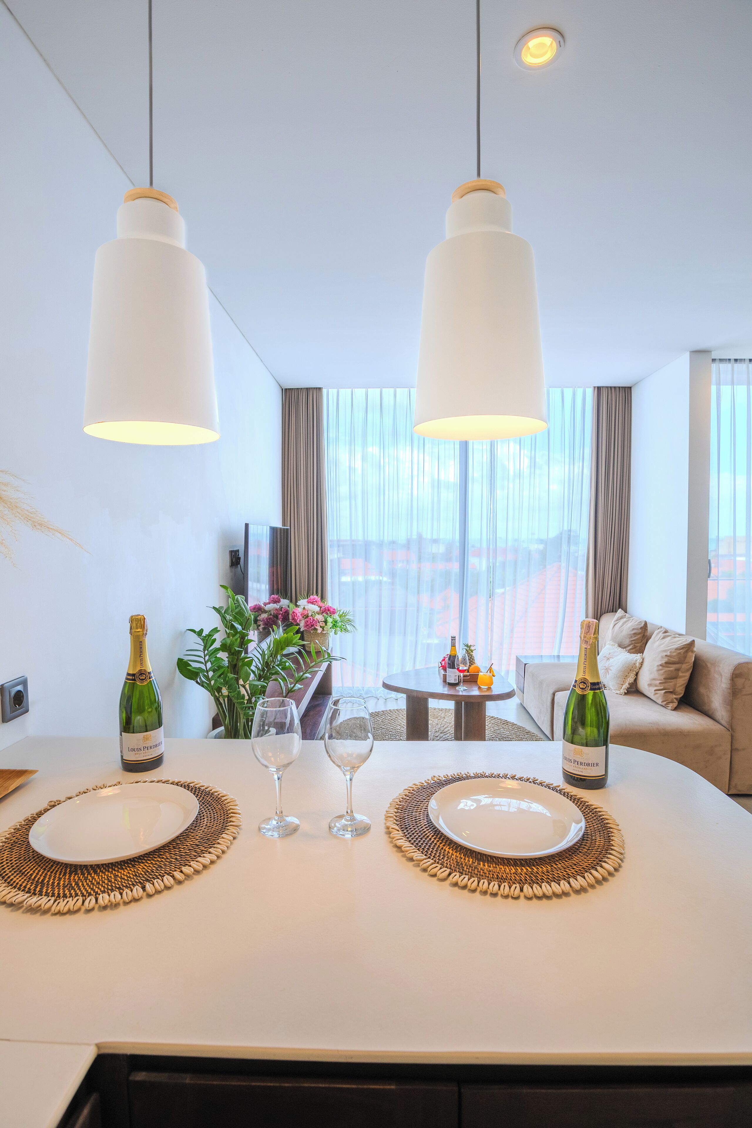 Un comedor moderno con una mesa blanca, dos copas de vino, dos botellas de vino y un jarrón de flores.