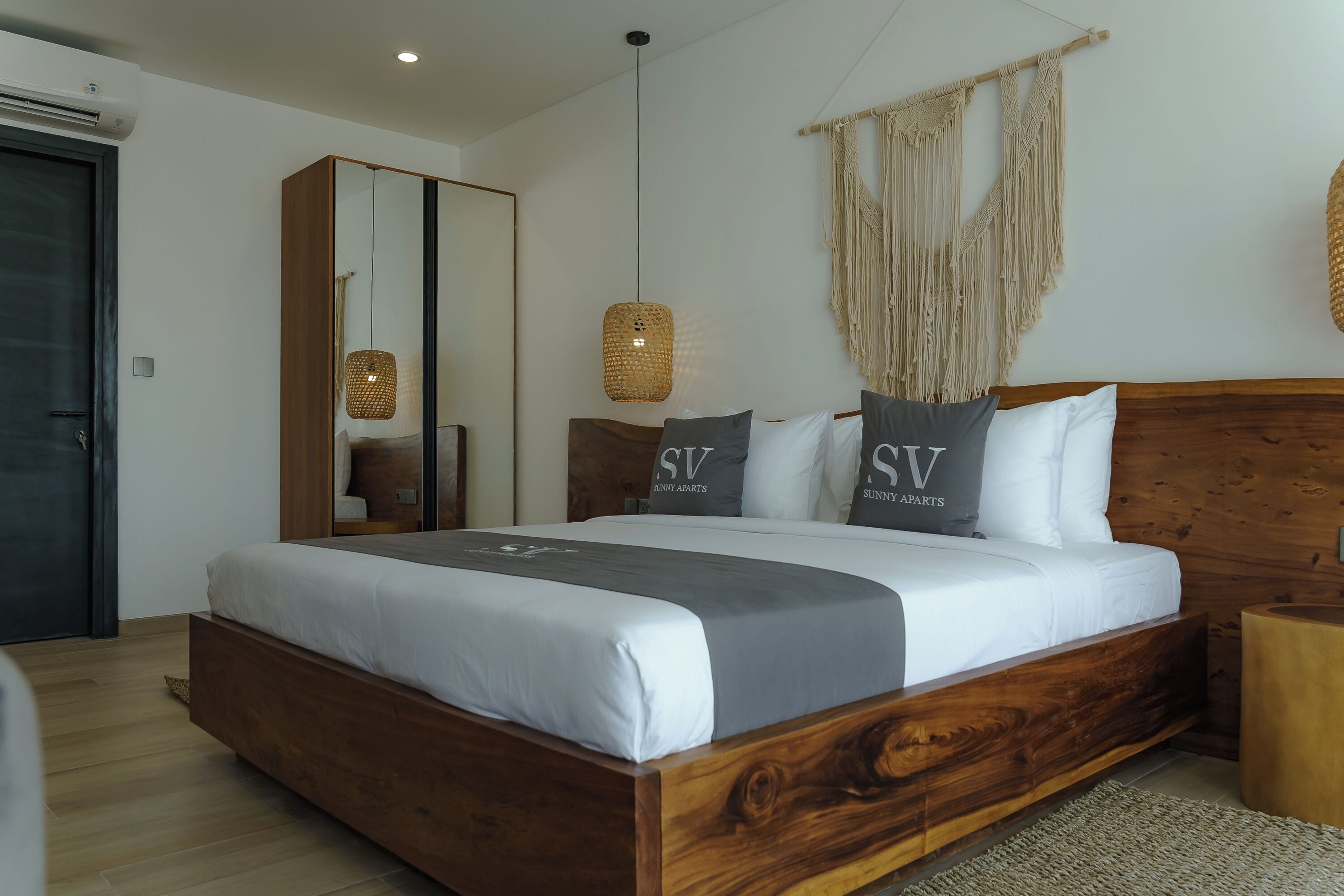 A modern bedroom with a wooden bed, a large mirror, macrame wall hanging, and a woven pendant light.
