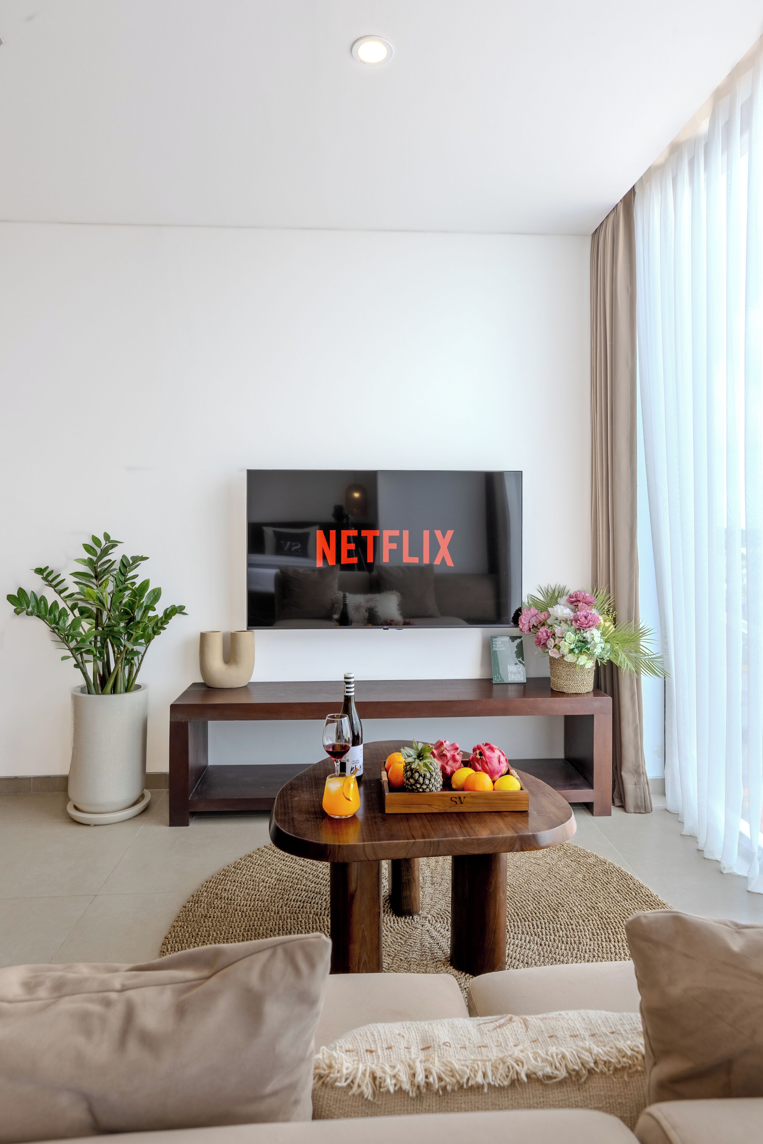 Un salon avec un téléviseur à écran plat affichant le logo « NETFLIX », une table basse en bois sur laquelle se trouvent des fruits et une bouteille, une plante en pot et un canapé avec des coussins.