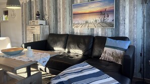 Un salon moderne avec un canapé en cuir noir, une table à manger en bois sur laquelle se trouvent un bol et une tasse blanche, et un grand tableau représentant un coucher de soleil sur l’eau, accroché au mur.