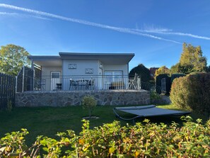 A modern two-story house with a stone retaining wall, a patio with outdoor furniture, and a trampoline in the yard. - Striking Holiday Home in Sternberg With Jetty (Sternberg)