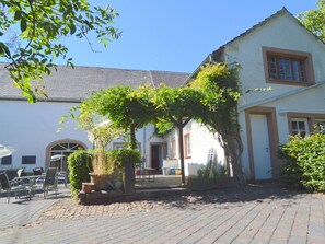Une cour avec un bâtiment blanc, une pergola couverte de plantes grimpantes, des sièges d’extérieur et un chemin pavé.