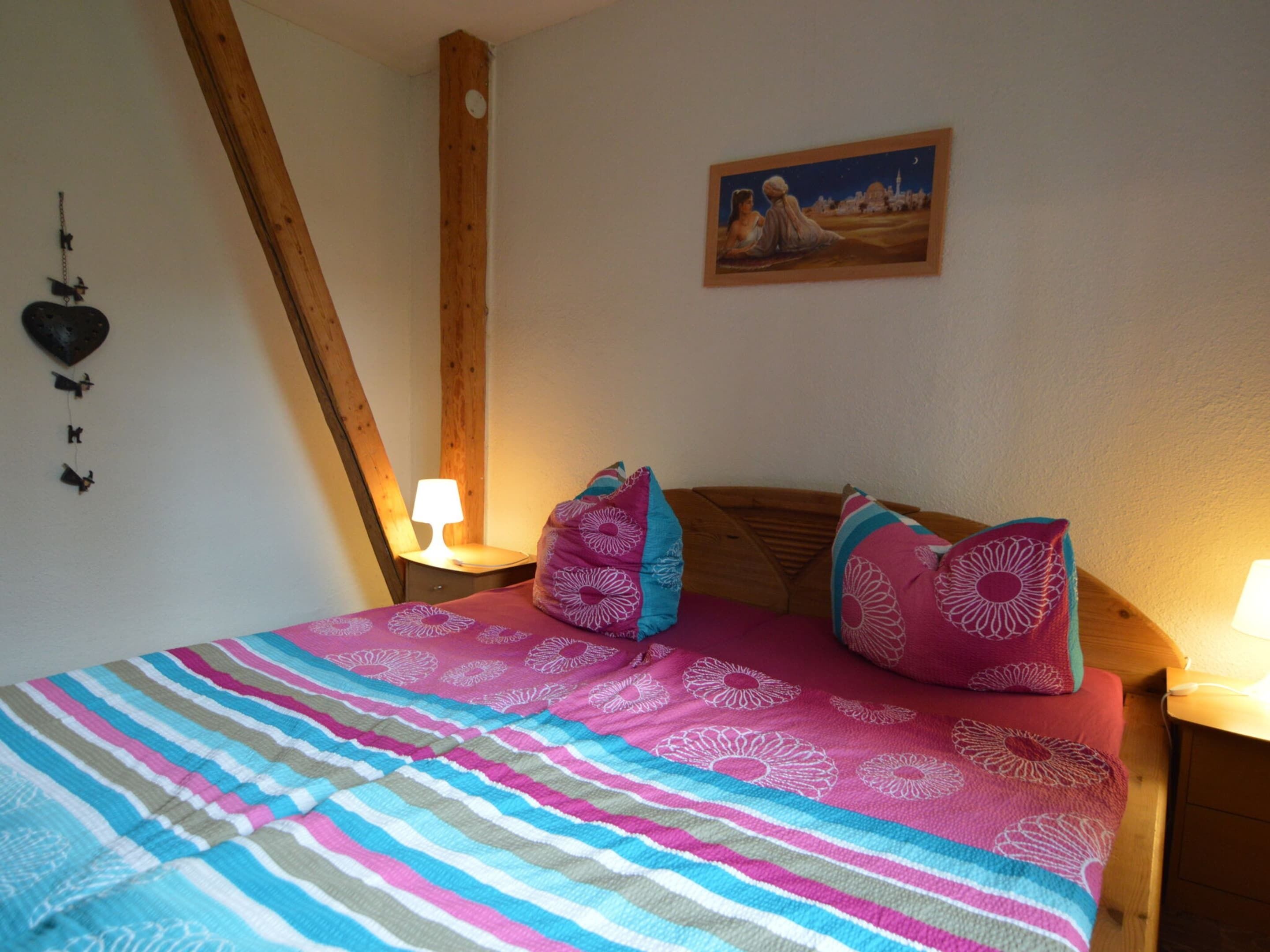 Un dormitorio con cama, almohadas, una mesita de noche, una lámpara de pared y un cuadro.