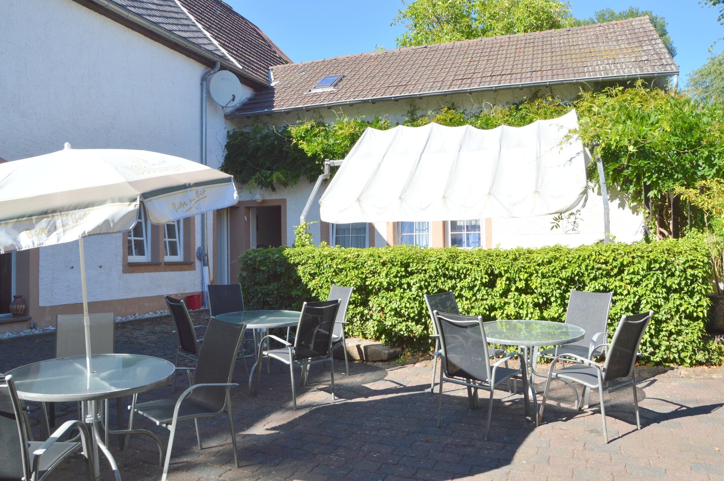 An outdoor patio with round tables and chairs, a white umbrella, and a building with a tiled roof and a hedge in the background.
