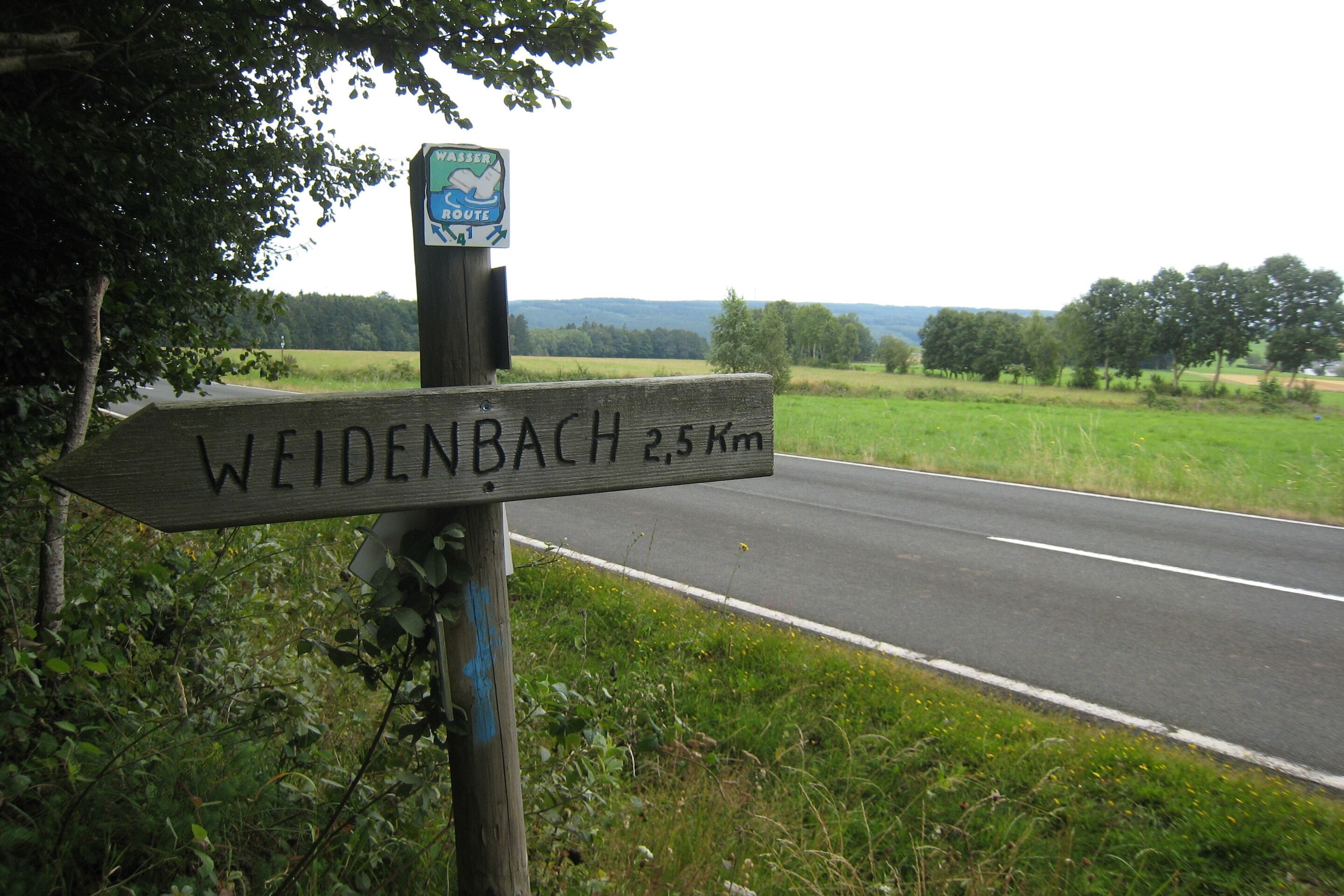 Un cartel de madera con la inscripción "WEIDENBACH 2.5 km" y un letrero que indica "RUTA DEL AGUA".