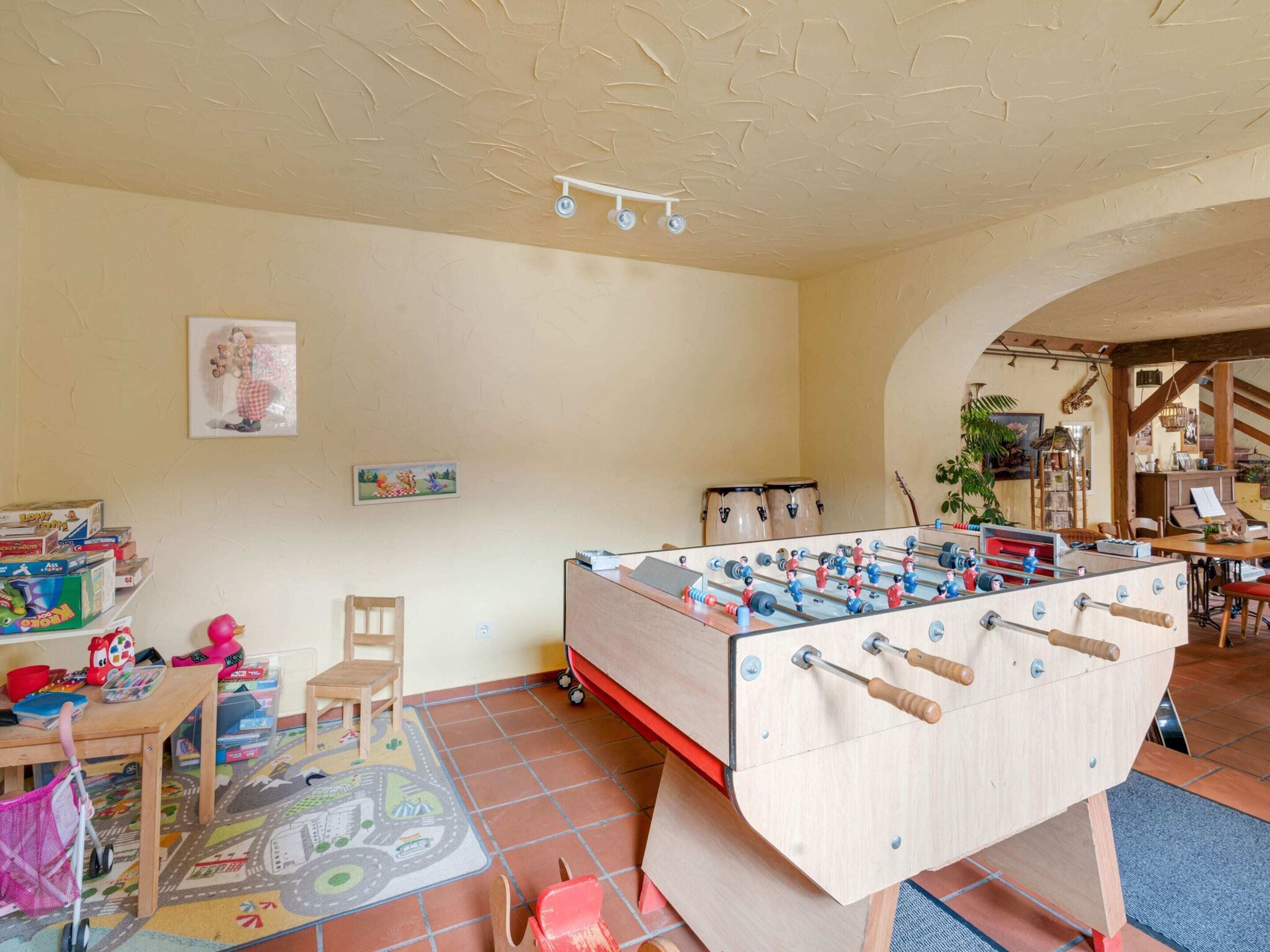 A cozy room with a foosball table, a small desk, and a chair. There are shelves with toys and books, and a colorful rug on the floor.