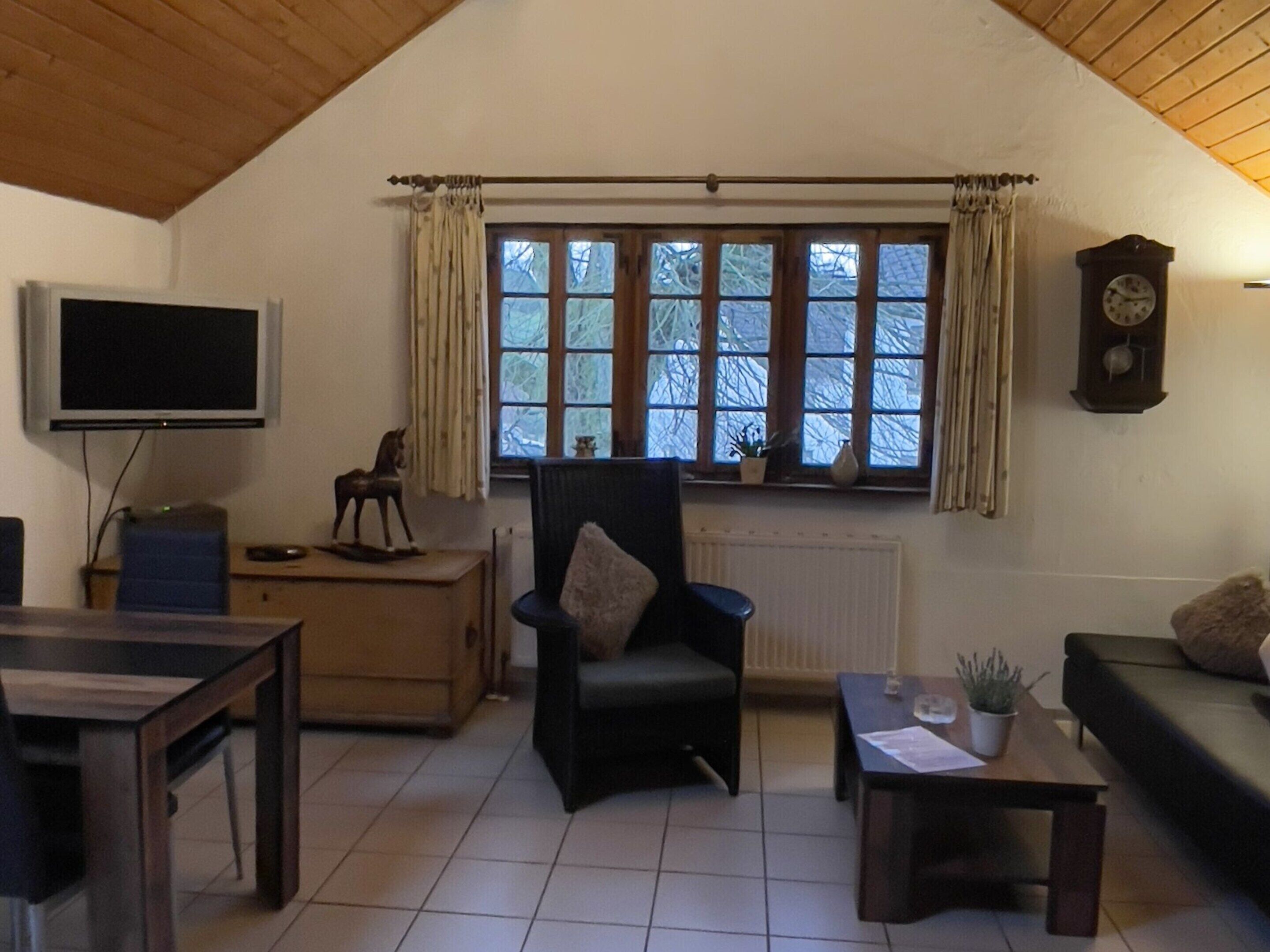 Un salon confortable avec une télévision, un meuble en bois, un fauteuil, un canapé et une fenêtre avec des rideaux.