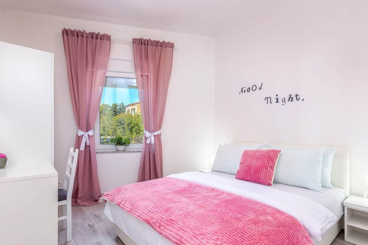 A bedroom with a bed, a window with curtains, a bedside table, and a wall with a motivational quote.