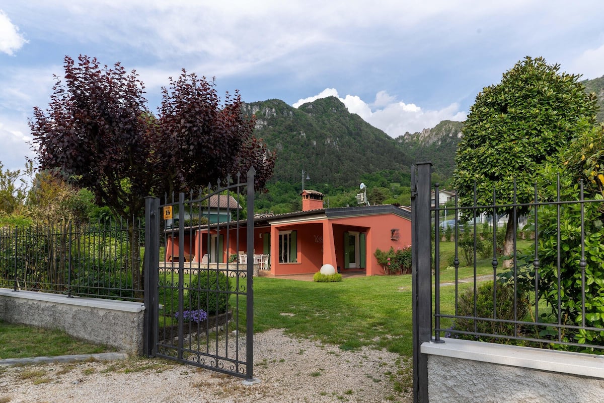 A red villa with a fenced garden, a mountain backdrop, and a gravel driveway.