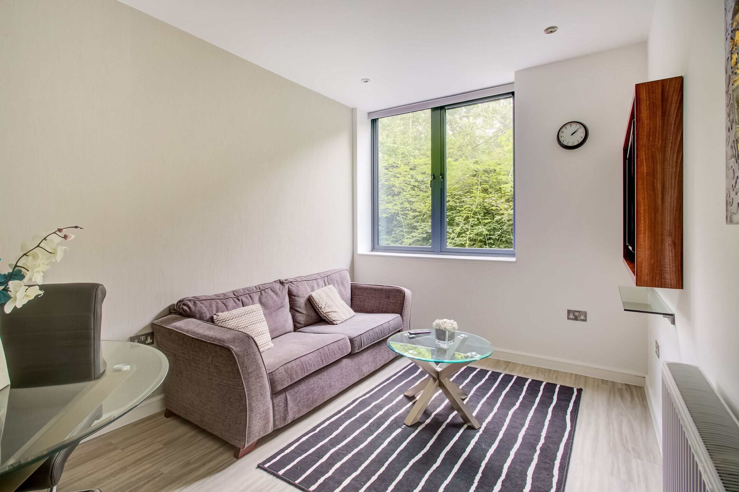 Een moderne woonkamer met een glazen salontafel, een bank, een stoel en een groot raam met uitzicht op groen.