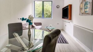 A modern living room with a glass dining table, a sofa, a TV mounted on the wall, and a window with a view of greenery. - Simplistic Apartment in Milton Keynes Near Willen Lake (Milton Keynes)