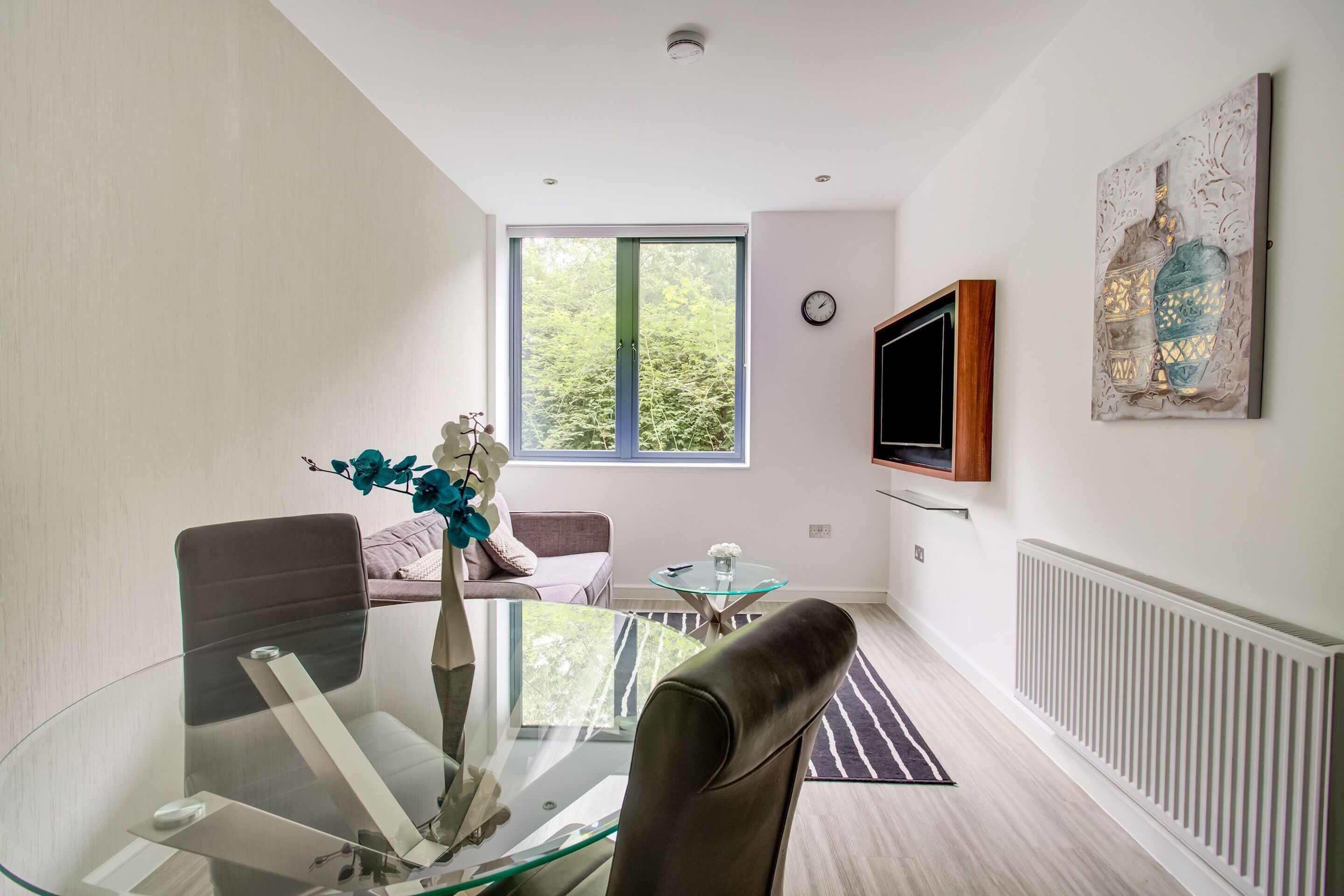 A modern living room with a glass dining table, leather chairs, a sofa, a TV mounted on the wall, and a large window with a view of trees.