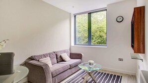 A modern living room with a sofa, a glass coffee table, a striped rug, a window with a view of greenery, and a clock on the wall. - Simplistic Apartment in Milton Keynes Near Willen Lake (Milton Keynes)