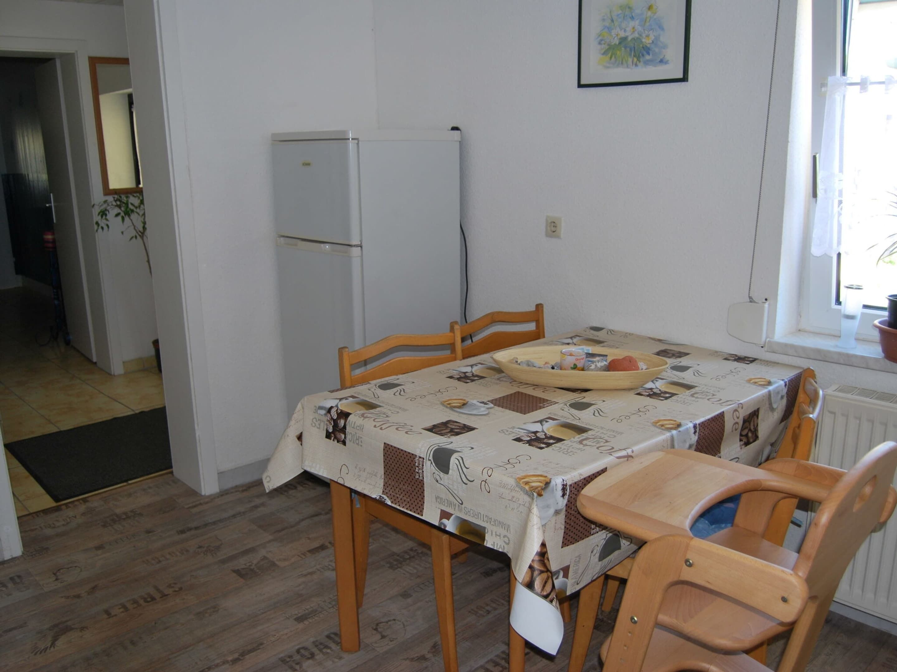 Ein Essbereich mit Tisch, bedeckt mit einer gemusterten Tischdecke, Holzsesseln, einer Schüssel mit Essen und einem weißen Kühlschrank.