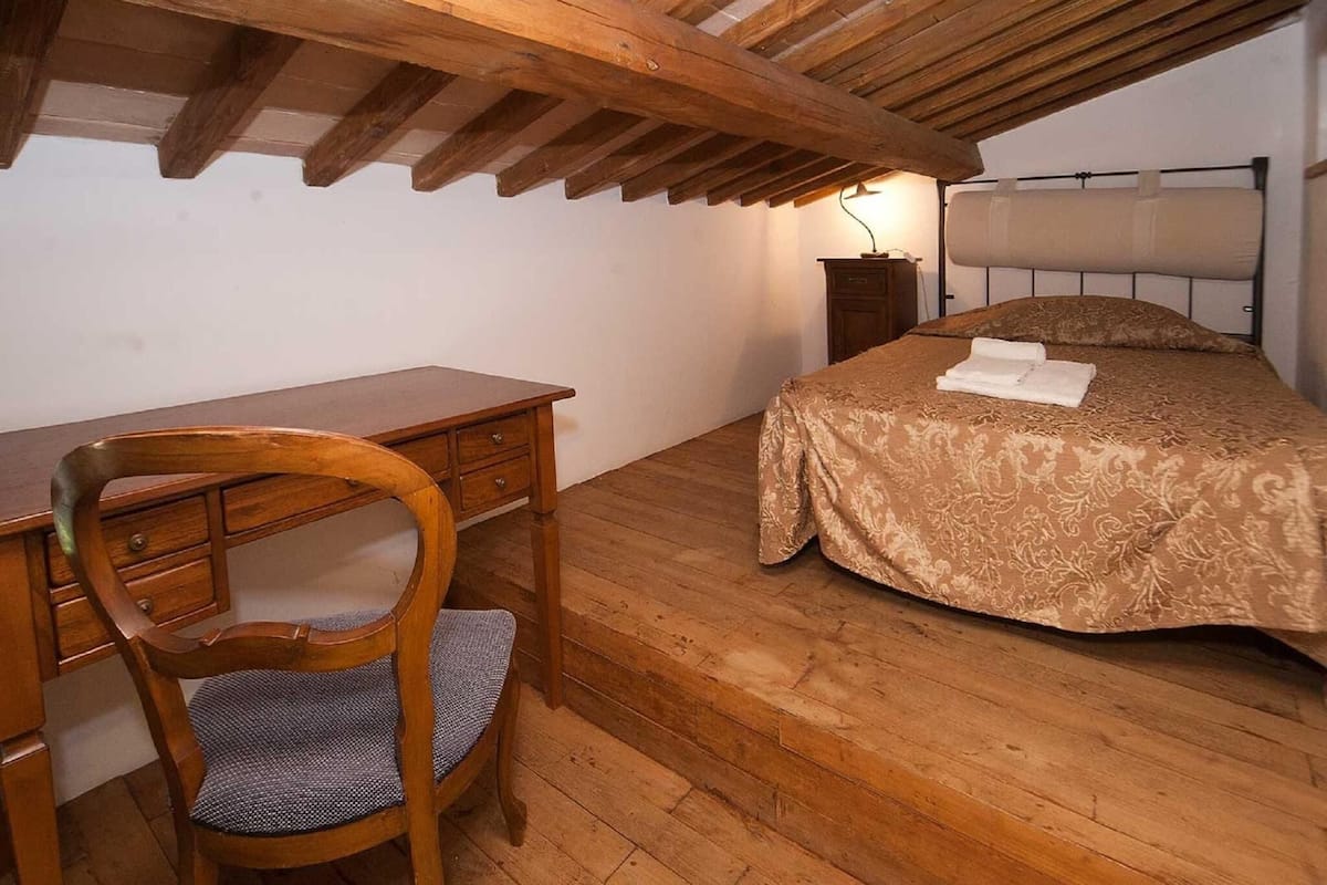 Un dormitorio con un escritorio de madera, una silla, una cama con colcha estampada y una mesita de noche con lámpara.