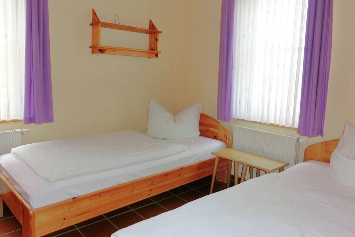 Una habitación con dos camas de madera, una mesita pequeña y un ventanal con cortinas moradas.