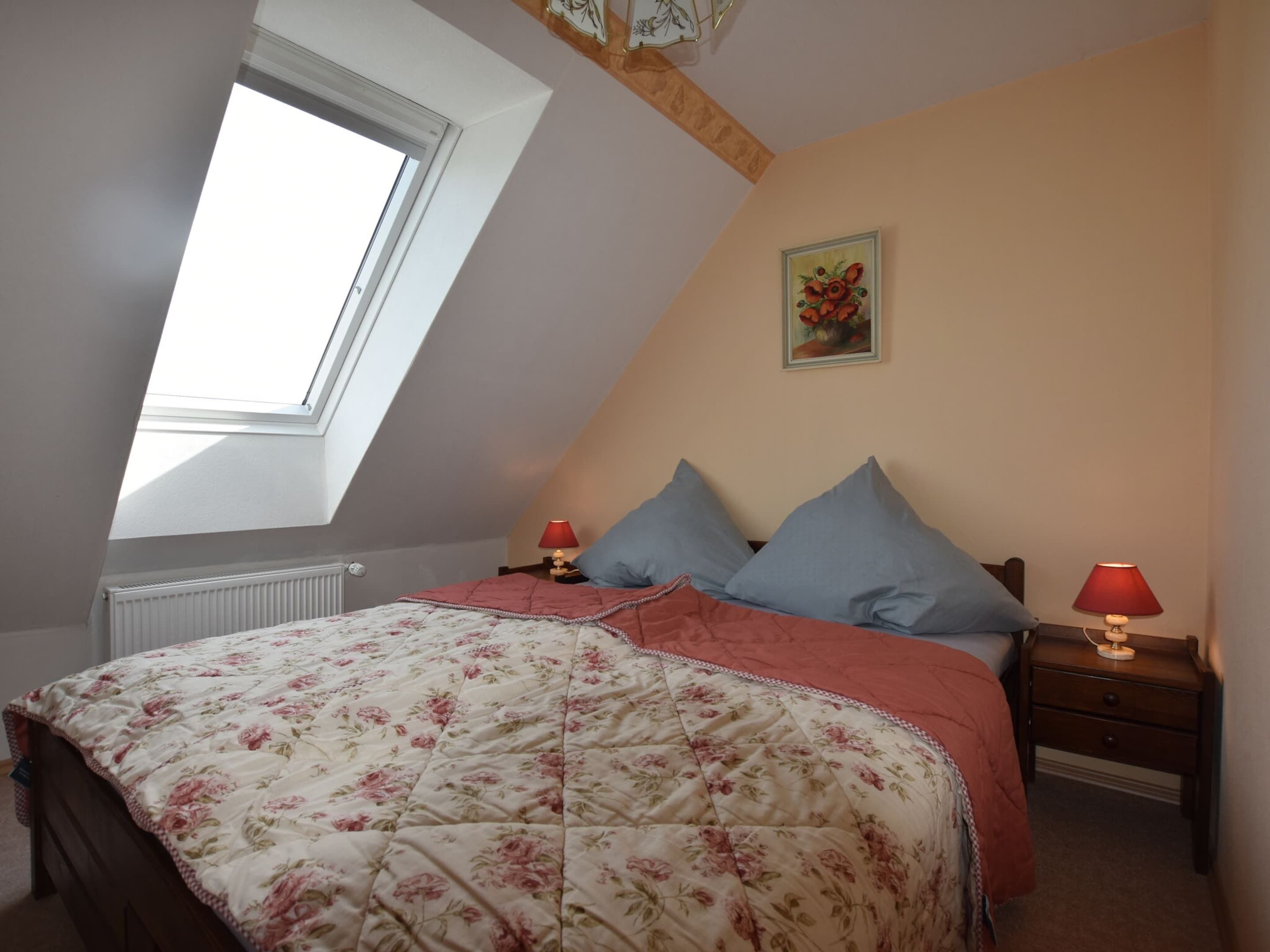 A bedroom with a sloped ceiling, a skylight, a floral bedspread, two bedside tables with lamps, and a painting on the wall.