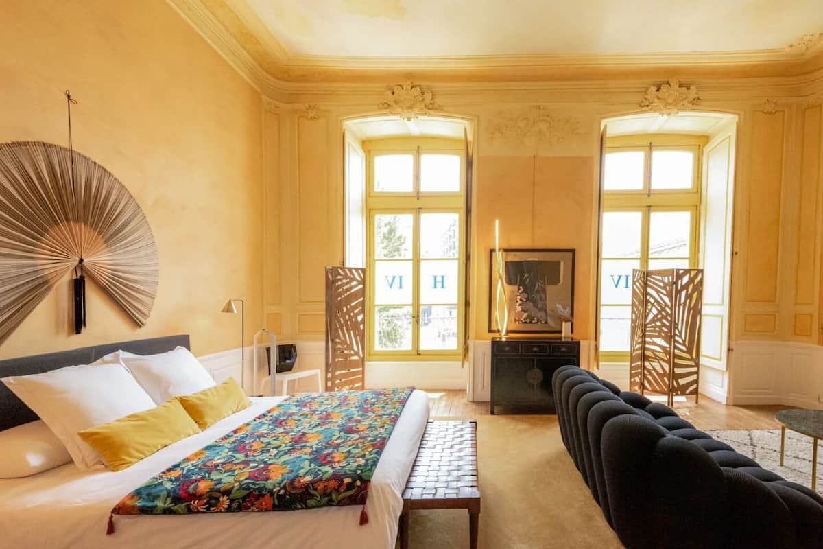 A hotel room with a bed, a desk, a chair, a fan, and two windows with curtains.