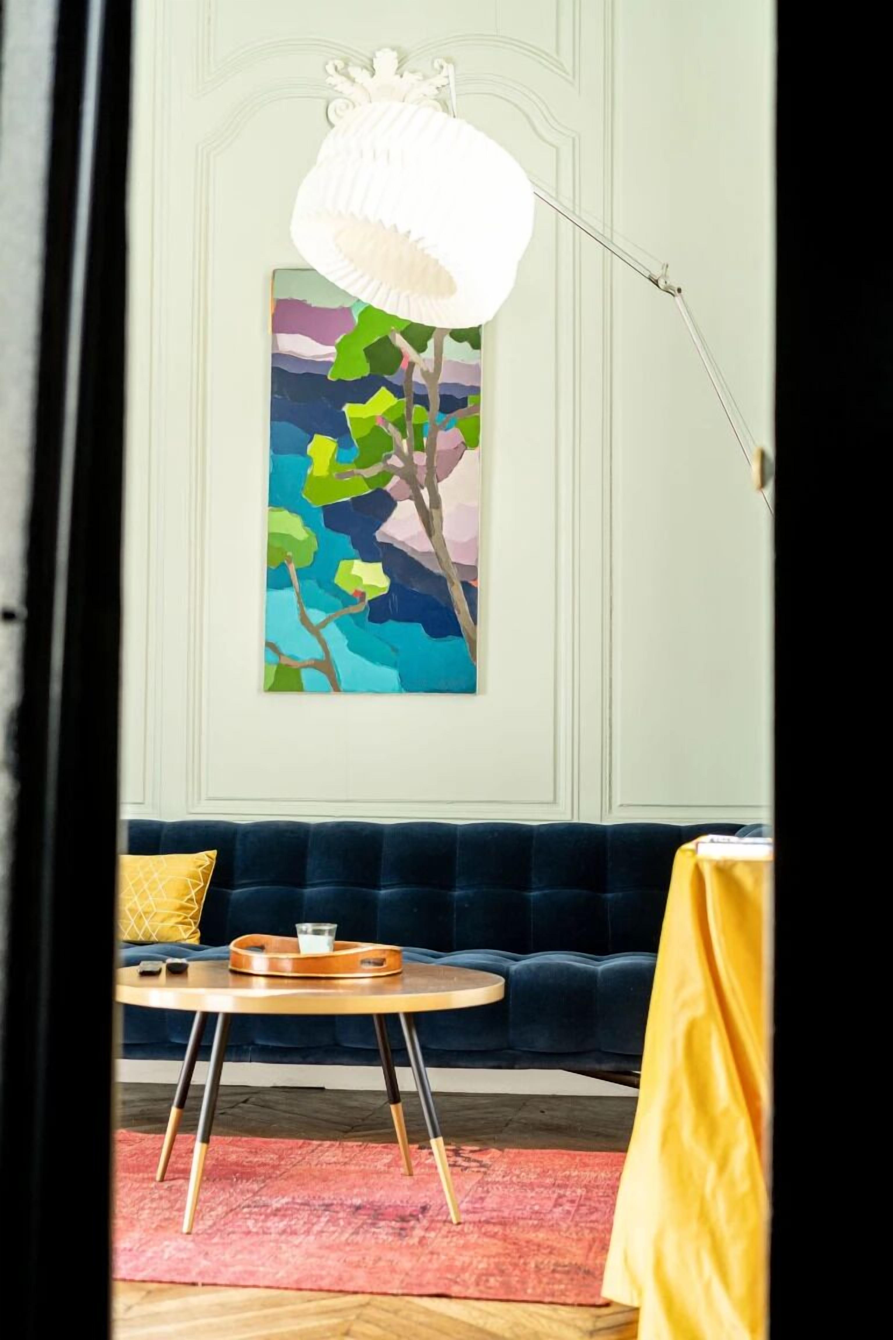 Ein Zimmer mit einem dunkelblauen Sofa, einem runden Holztisch, einem farbenfrohen abstrakten Gemälde an der Wand und einer weißen Stehlampe.