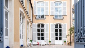 Une cour intérieure avec un bâtiment doté de volets blancs, une petite table avec des chaises et des plantes en pot.