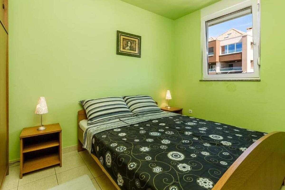 A small, single-bed room with a green wall, a framed picture, a window with a view of buildings, and two bedside lamps.