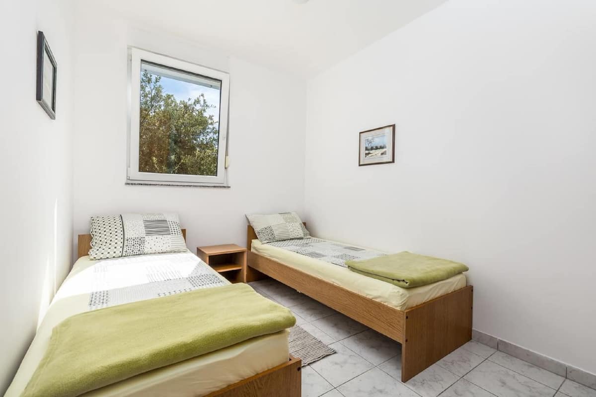 A small, clean room with two single beds, a window with a view of trees, and a small bedside table.