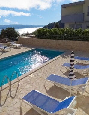 A swimming pool with lounge chairs and an umbrella, surrounded by a stone deck and a building with a view of the sea. - Charming Apartment in Makarska With Pool (Makarska)