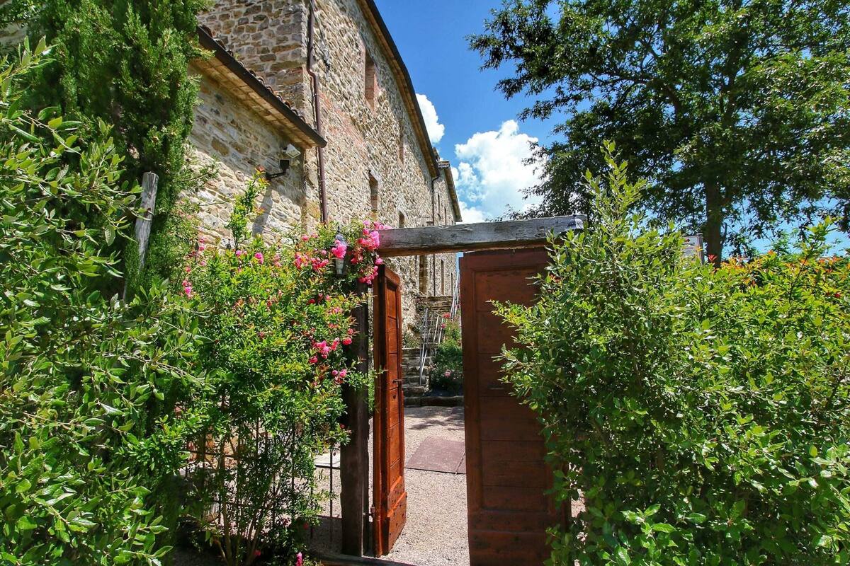 Un edificio de piedra con una puerta de madera, rodeado de abundante vegetación y plantas en flor.
