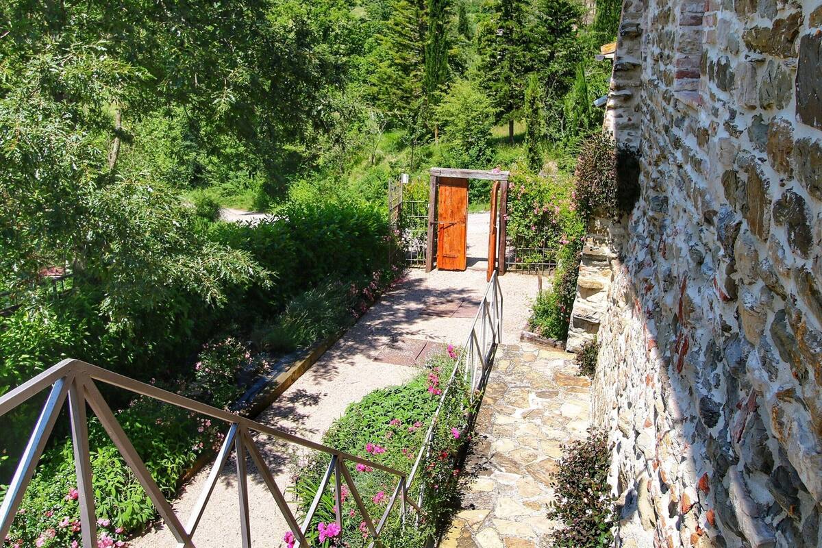 Un sendero de piedra que conduce a una puerta de madera, rodeado de abundante vegetación y un muro de piedra.